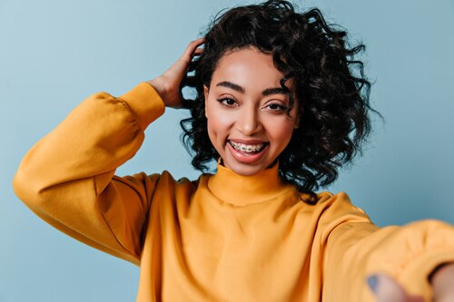 Mulher jovem com cabelos cacheados sorri com confiança usando aparelho nos dentes. Ela veste amarelo e está sobre um fundo azul