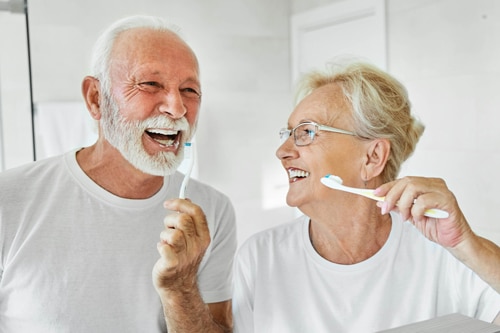 Homem e mulher idosos sorrindo enquanto escovam os dentes, representando a importância da prevenção odontológica ao longo da vida.