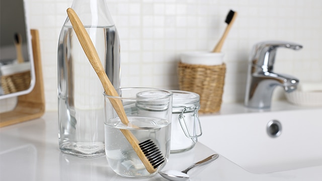 Escova sendo limpa com solução de vinagre, água e bicarbonato sobre a pia do banheiro, em ambiente limpo e natural.