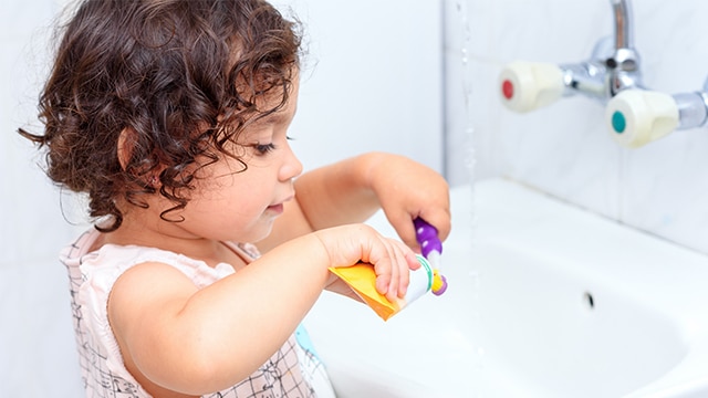 Criança pequena em frente a uma pia, colocando creme dental sem flúor na escova de dentes