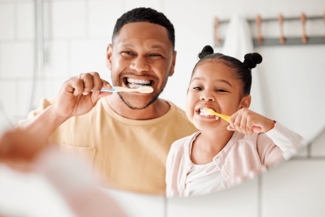 Criança escovando dente ao lado do pai e se divertindo enquanto cuida da sua saúde bucal.
