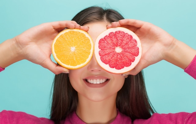 Mulher sorridente segura frutas ácidas nos olhos, com uma rodela de laranja e outra de toranja, em um fundo azul claro.