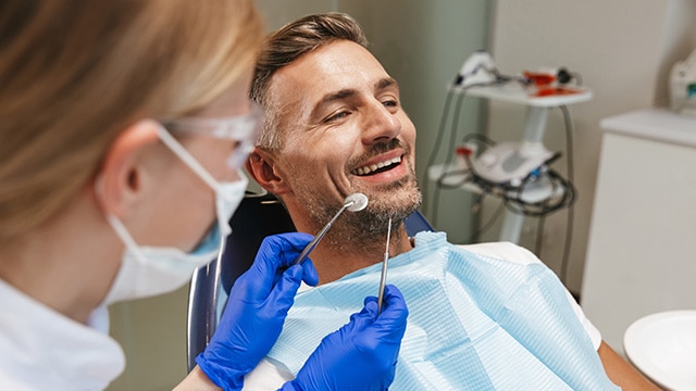 Homem adulto sorri relaxado durante consulta odontológica enquanto dentista, usando luvas azuis e máscara, segura instrumentos de exame próximos à sua boca.