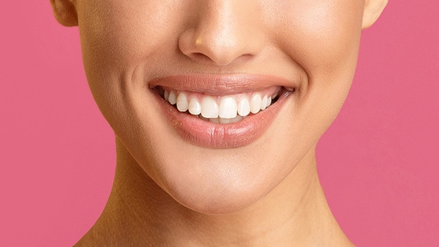 Close do sorriso de uma pessoa com dentes brancos e perfeitamente alinhados sobre um fundo rosa sólido.