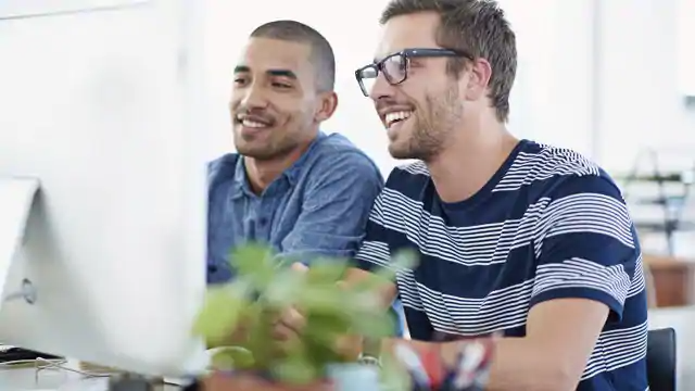 dois homens, sendo um moreno de camisa azul e um loiro de óculos e camiseta listrada, sentados em frente a um computador e sorridentes.