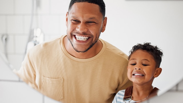 Pai e filho sorrindo em frente ao espelho durante cuidados de saúde bucal no Dia Mundial da Saúde Gengival.