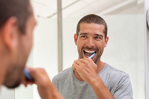 Homem em close-up utilizando escova de dentes azul, refletido no espelho. Foco na técnica de escovação e saúde bucal preventiva.