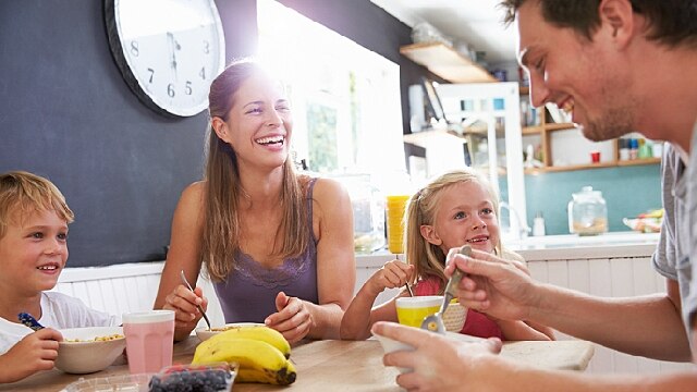 quatro pessoas sentadas em uma mesa: dois adultos e duas crianças. À direita, um homem de cabelo escuro está sorrindo, enquanto come algo que está em uma tigela. Ao seu lado, uma menina de cabelos nos ombros e loiros sorri. Ao lado, uma mulher de cabelos longos e claros sorri. Na outra ponta da mesa, um menino de cabelos curtos e claros também  sorri.