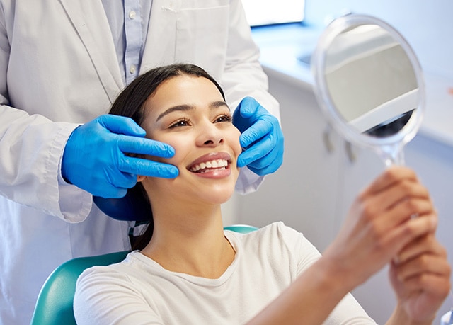 Mulher sorrindo e segurando espelho para observar a lente de resina aplicada por dentista em consultório odontológico.