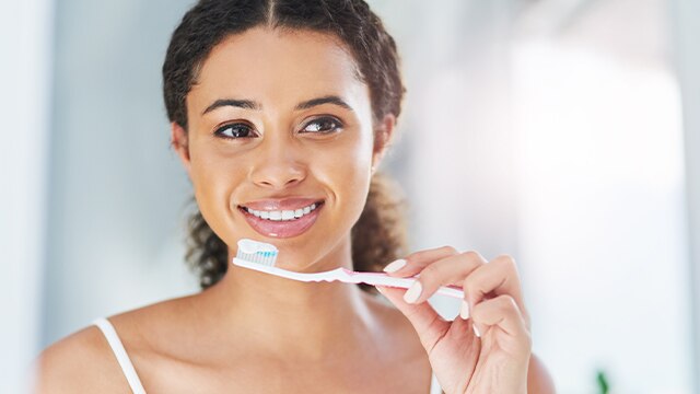 Mulher de camiseta branca em frente ao espelho sorrindo ao mostrar como escovar os dentes