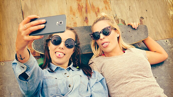 Amigas deitadas no chão com as cabeças apoiadas em um skate tirando uma selfie