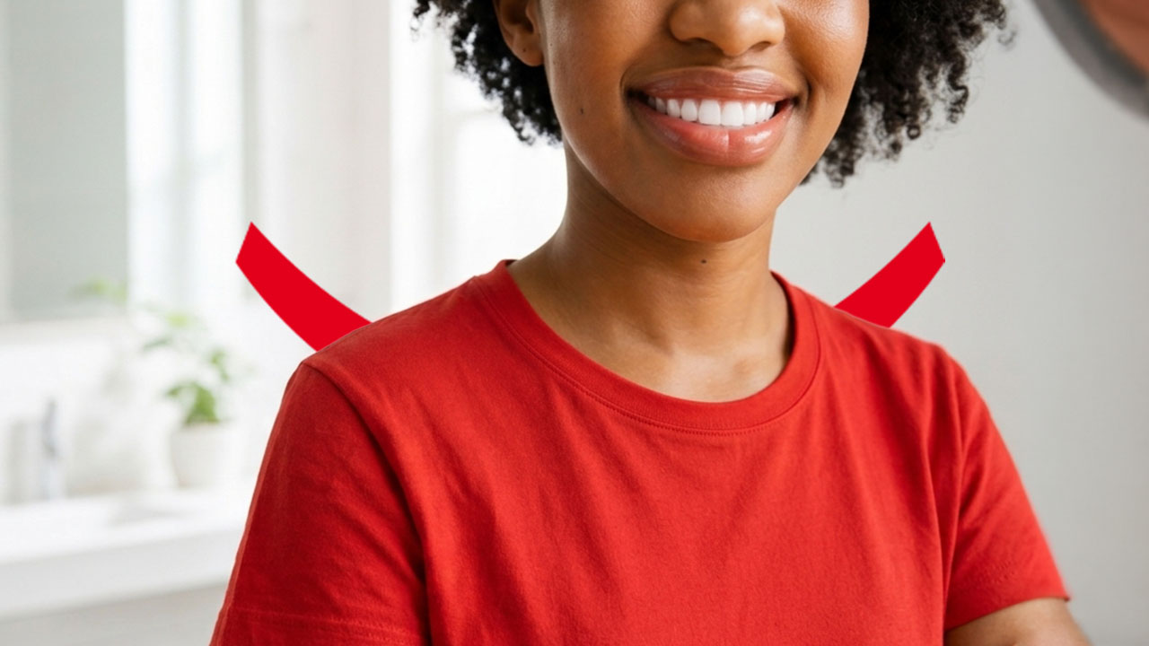 Uma mulher sorridente, com cabelos escuros e cacheados e uma camiseta vermelha, em pé em um banheiro todo branco.
