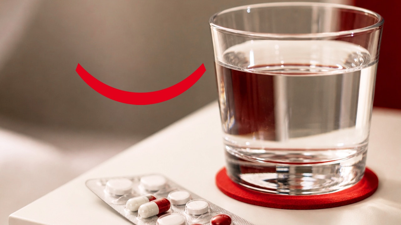 Um copo de água na mesinha de cabeceira, com um porta-copos vermelho ao lado de uma embalagem de comprimidos para azia e outros medicamentos.