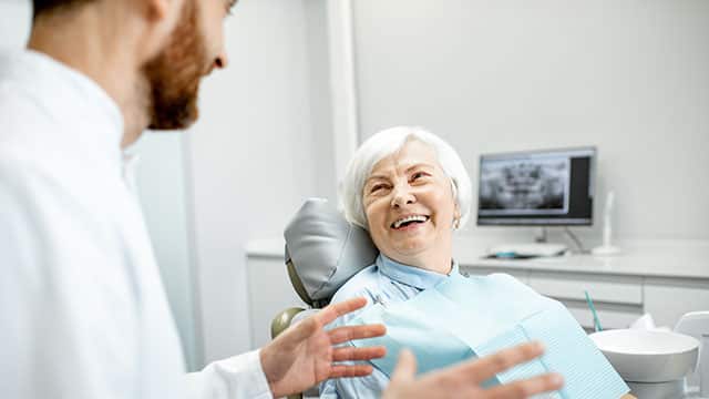 mulher idosa de cabelos brancos e curtos, sentada em uma cadeira odontológica, com roupas claras, sorrindo e esperando ser atendida pelo dentista, que está sentado ao seu lado. Ele é branco, de cabelos e barba castanhos e está sentado na frente dela