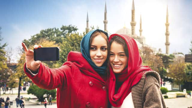 duas mulheres tirando uma selfie. A mulher à esquerda veste casaco vermelho e tem cabelos claros. A mulher à direita veste um casaco marrom com uma blusinha branca por baixo. Ambas estão sorrindo. 