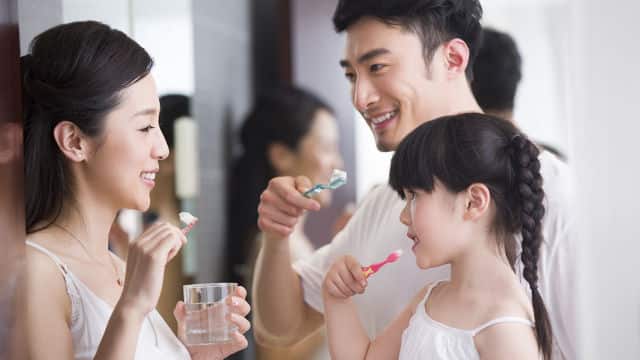 um homem jovem, uma mulher jovem e uma criança asiáticos com roupas brancas sorrindo e escovando os dentes