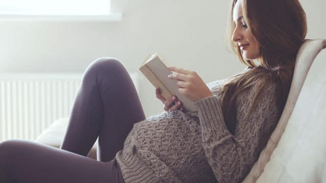Mulher gestante com cabelos longos e escuros, vestindo calça marrom e suéter cinza, lendo um livro no sofá.