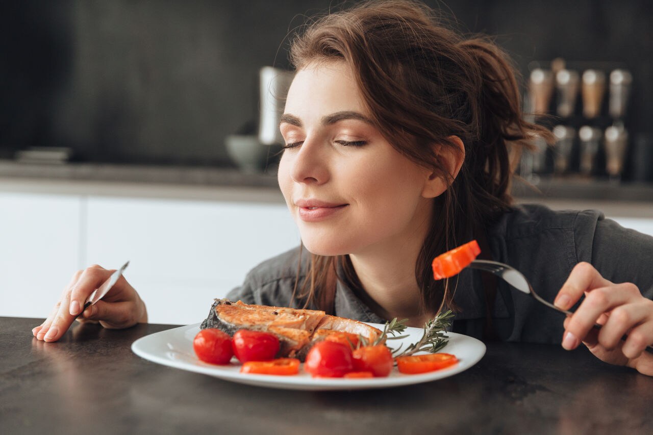 Image of pretty young woman sitting in kitchen while eating and smells fish and tomatoes. Eyes closed.; Shutterstock ID 579004342; Purchase Order (images grouped in same invoice by PO number): -; Client/Licensee: -