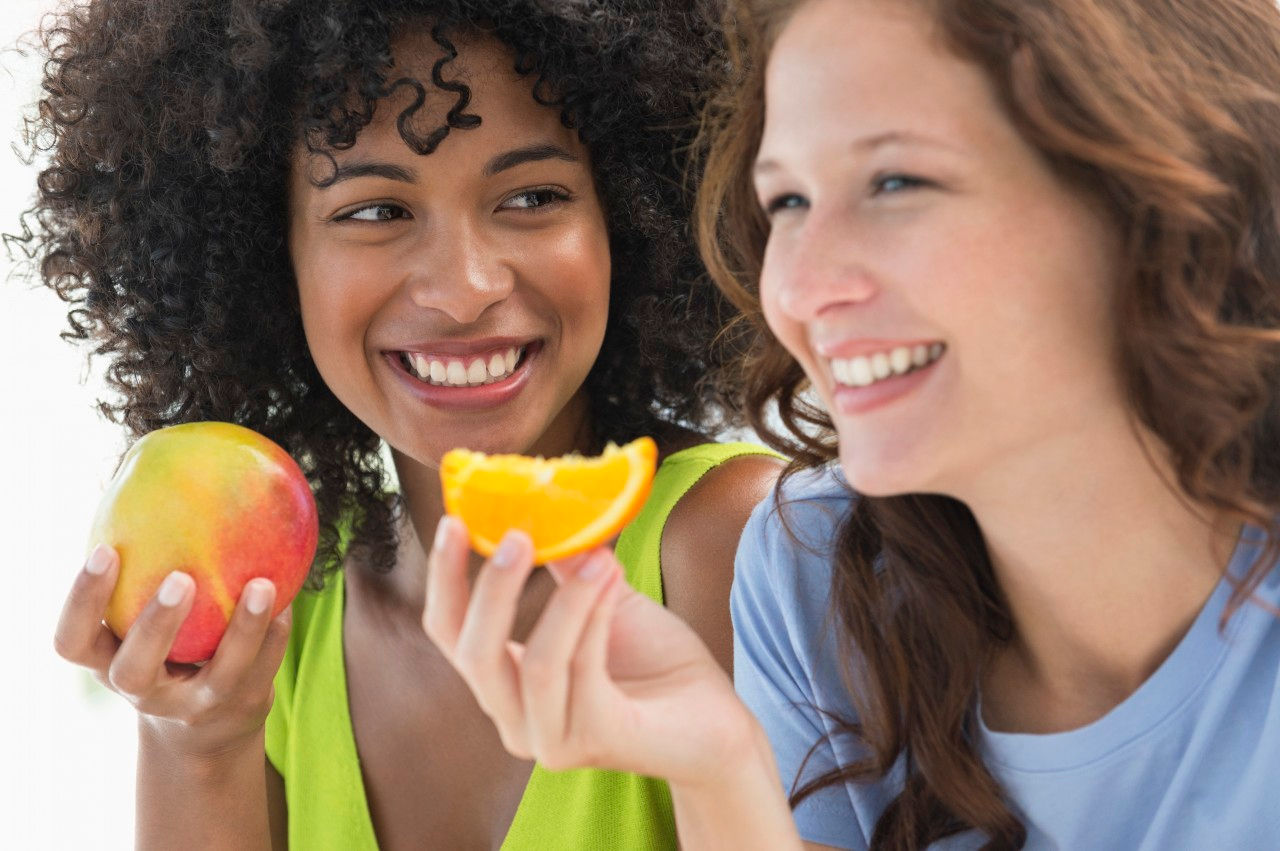 Duas mulheres sorridentes seguram frutas ácidas, como laranja e manga, enquanto conversam e se divertem juntas.