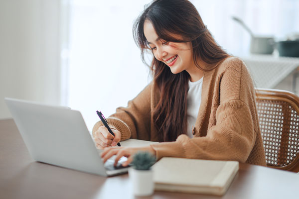 Mulher sentada em frente ao seu computador fazendo anotações de perguntas importantes para fazer durante sua entrevista de emprego