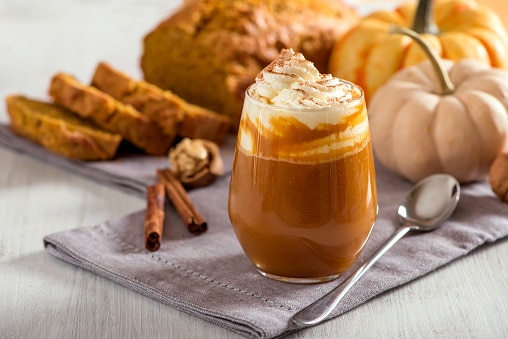 Copo de café cremoso com chantilly e canela, decorado em um ambiente aconchegante com pães e abóbora ao fundo.