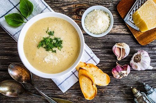 Tigela de sopa de legumes cremosa, acompanhada de pão, queijo ralado e temperos frescos sobre uma mesa rústica. 